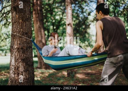 Una giovane coppia gode di un momento di relax in un'amaca sotto gli alberi, con una donna sorridente e un uomo in avvicinamento Foto Stock