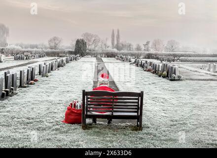 Una persona seduta da sola su una panchina di legno in un cimitero ricoperto di gelo, di nuovo alla macchina fotografica, vestita da Babbo Natale. REGNO UNITO Foto Stock