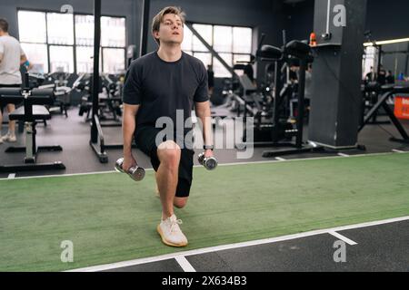 Ritratto di un giovane atleta atletico principiante motivato in abbigliamento sportivo che tiene i manubri che fanno affondi di squat e si riscalda allenandosi in palestra. Foto Stock
