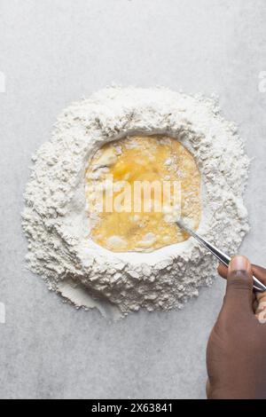 Vista dall'alto dell'impasto degli spaghetti all'uovo mescolato su un ripiano in marmo, mentre le uova e la farina vengono mescolate per preparare gli spaghetti all'uovo fatti in casa e la pasta all'uovo Foto Stock