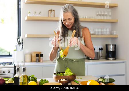 A casa, donna caucasica matura che indossa grembiule verde, che prepara l'insalata Foto Stock