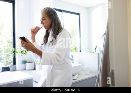 A casa, donna caucasica matura che pulisce i denti, controllando lo smartphone Foto Stock