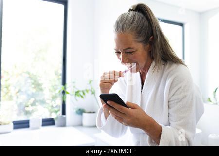 A casa, donna caucasica matura che si lava i denti, controllando lo smartphone Foto Stock