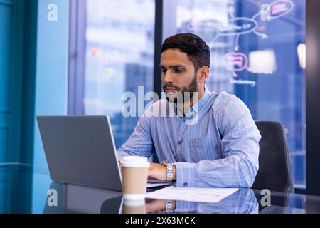 In ufficio, un professionista birazziale maschile che si concentra sul laptop, tenendo documenti aziendali Foto Stock