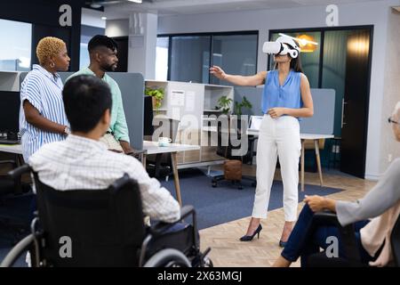 In ufficio, un team eterogeneo che ascolta donne caucasiche con il visore VR che punta Foto Stock
