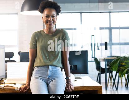 In ufficio, donna d'affari afroamericana seduta sulla scrivania, sorridente alla macchina fotografica Foto Stock