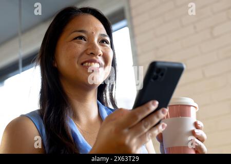 In ufficio, donna d'affari birazziale che tiene lo smartphone, guardando lontano Foto Stock