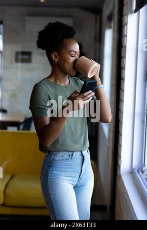 In ufficio, donna d'affari afroamericana che beve caffè, controlla il telefono Foto Stock