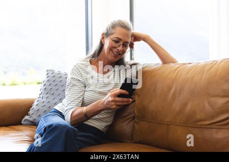A casa, donna caucasica matura che indossa gli occhiali che controlla il suo telefono sul divano Foto Stock