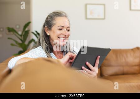 A casa, donna caucasica matura che tiene un tablet, ridendo Foto Stock