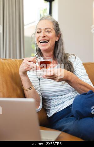 A casa, una donna caucasica matura che tiene il tè, ridendo del portatile Foto Stock