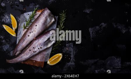 Nasello di pesce crudo fresco guarnito con fette di rosmarino e limone su una superficie scura, perfetto per temi culinari o concetti alimentari sani. Extra Foto Stock