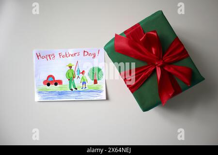 Festa del papà foto di una cartolina disegnata da un bambino. Un regalo di colore verde con un fiocco rosso. Sfondo bianco. La cartolina mostra padre e figlio a pescare. Foto Stock