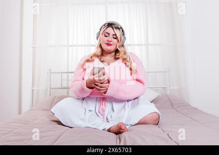 Donna allegra e di grandi dimensioni seduta su un letto, che ascolta musica con le cuffie e utilizza il suo smartphone Foto Stock