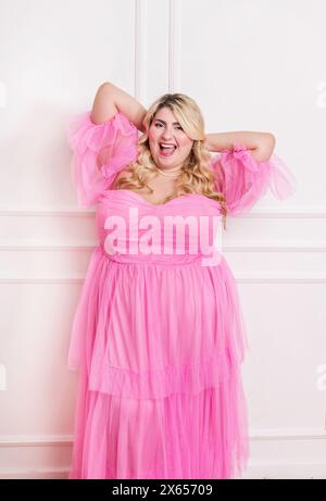 Donna radiosa in versione Plus size che posa in studio con entusiasmo in un elegante abito rosa Foto Stock