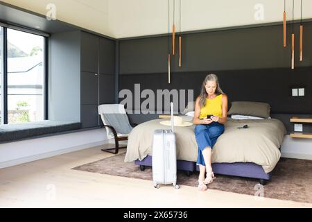 A casa, donna caucasica matura seduta sul letto, guardando lo smartphone Foto Stock