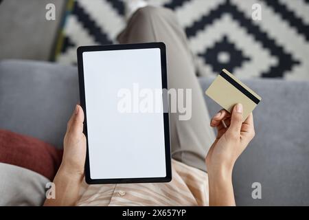 Dall'alto della vista di una donna irriconoscibile seduta sul divano in salotto con un tablet digitale che paga online con carta di credito Foto Stock