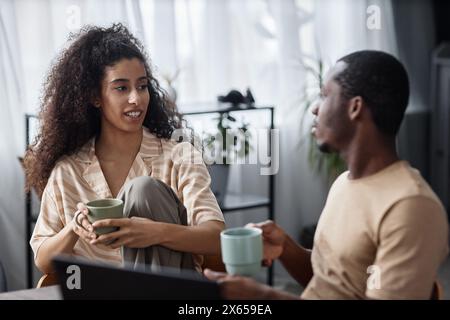 Giovane donna birazziale e il suo ragazzo afroamericano che tiene tazze con tè o caffè seduti in salotto e che parla Foto Stock