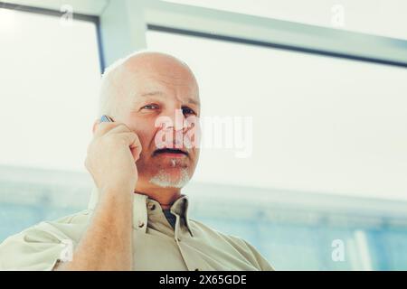 Un professionista senior si impegna attivamente in una conversazione telefonica, davanti a uno sfondo architettonico contemporaneo, vestito con abbigliamento casual per affari Foto Stock