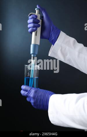 Analisi di laboratorio. Scienziato che gocciola il campione con la micropipetta nel becher su sfondo scuro, primo piano Foto Stock