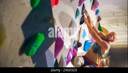 Forte scalatore esperto che pratica l'arrampicata in solitaria sul muro di Boulder in palestra. Uomo che fa esercizio presso la palestra Indoor fitness Facility, praticando sport estremi per il suo sano allenamento Lifestyle. Primo piano verticale Foto Stock