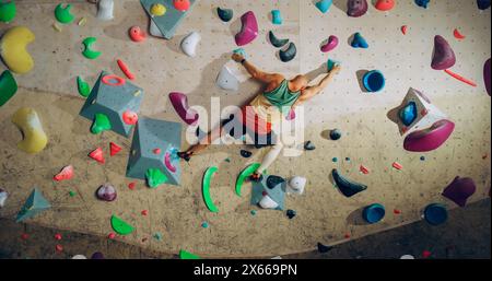 Forte esperto arrampicatore che pratica arrampicata in solitaria sul muro di Bouldering in una palestra. Uomo che fa esercizio presso la palestra Indoor fitness Facility, praticando sport estremi per il suo sano allenamento Lifestyle. Sparato da dietro. Foto Stock
