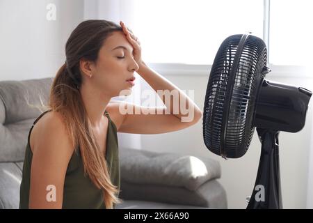 Una giovane donna surriscaldata davanti al ventilatore che lavora soffre di caldo estivo a casa. Ragazza seduta di fronte al ventilatore quando fa caldo. Estate calda Foto Stock