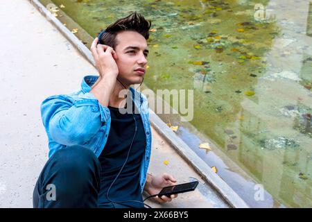 Il giovane con le cuffie usa lo smartphone vicino a un laghetto, indossando una camicia in denim e pantaloni neri. Si siede concentrato, in un ambiente urbano rilassato all'aperto. Foto Stock