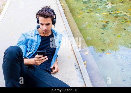 Il giovane con le cuffie usa lo smartphone vicino a un laghetto, indossando una camicia in denim e pantaloni neri. Si siede concentrato, in un ambiente urbano rilassato all'aperto. Foto Stock