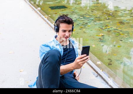 Il giovane con le cuffie usa lo smartphone vicino a un laghetto, indossando una camicia in denim e pantaloni neri. Si siede concentrato, in un ambiente urbano rilassato all'aperto. Foto Stock