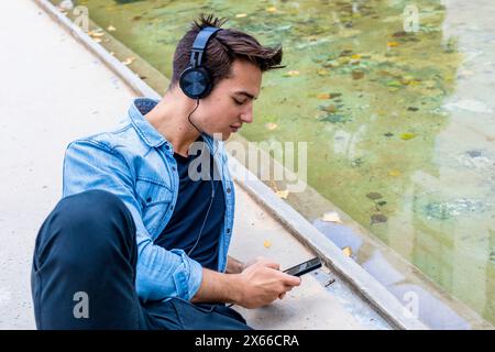 Il giovane con le cuffie usa lo smartphone vicino a un laghetto, indossando una camicia in denim e pantaloni neri. Si siede concentrato, in un ambiente urbano rilassato all'aperto. Foto Stock