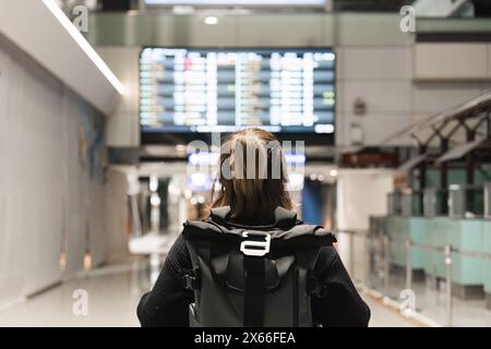 Giovane donna zaino nel terminal dell'aeroporto internazionale che guarda il tabellone informazioni sugli orari dei voli e controlla il suo volo Foto Stock