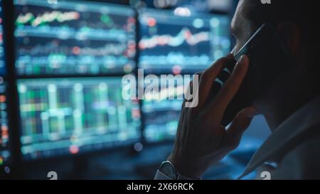 Financial Day Trader che lavora su un computer con workstation multi-monitor con grafici delle azioni in tempo reale e parla al telefono con un cliente, mentre si trova in ufficio a tarda serata. Primo piano. Foto Stock