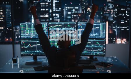 Il broker del mercato azionario che lavora su un computer con workstation multi-monitor, vince e celebra il successo alzando le mani in alto. L'uomo d'affari lavora sull'analisi dei dati in Office at Night. Foto Stock