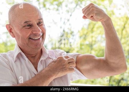 L'uomo di 65 anni che rimane in forma, anziano carismatico maturo flette i muscoli del braccio, mostrando forza, corpo sano, mente sana Foto Stock