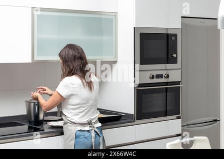 Una persona in una cucina moderna che cucina con una pentola sul piano cottura, indossa un top bianco e jeans blu Foto Stock