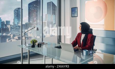 Ritratto di un'innovativa donna d'affari musulmana che indossa Burka seduto alla sua scrivania che lavora su un computer portatile nel Big City Office. Strategia di investimento per il successo del piano del CEO aziendale per l'avvio dell'e-Commerce Foto Stock