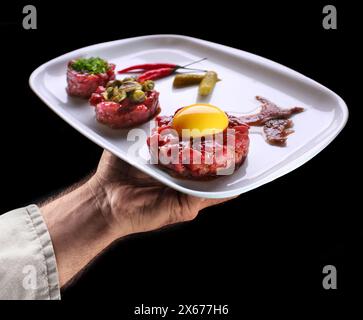 Lo chef tiene un piatto bianco di tartare di bistecca su sfondo nero. Cibo gustoso. Foto Stock