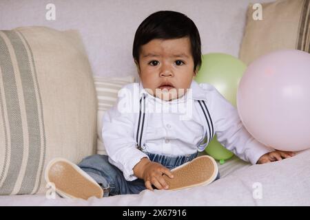 Ritratto di un simpatico bambino seduto su un divano, vestito con un'elegante camicia bianca con bretelle e jeans in denim. Foto Stock