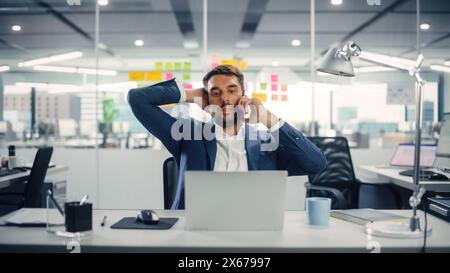 Giovane uomo d'affari felice che usa il computer e parla al telefono nell'ufficio moderno. Bel manager di successo sorridente, che lavora a progetti finanziari e di marketing. Bere tè o caffè da una tazza. Foto Stock