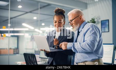 Senior Manager che parla con un uomo d'affari esperto e sicuro mentre utilizza un computer portatile in ufficio. I colleghi discutono di progetti commerciali, finanziari e di marketing. Gli specialisti lavorano in team diversificati. Foto Stock