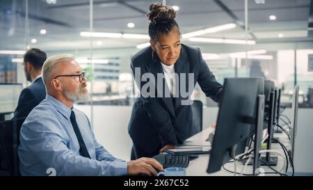 Senior Manager che parla con un uomo d'affari esperto e sicuro di sé mentre utilizza il computer in ufficio moderno. I colleghi discutono di progetti commerciali, finanziari e di marketing. Gli specialisti lavorano in team diversificati. Foto Stock