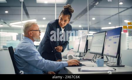 Senior Manager che parla con un uomo d'affari esperto e sicuro di sé mentre utilizza il computer in ufficio moderno. I colleghi discutono di progetti commerciali, finanziari e di marketing. Gli specialisti lavorano in team diversificati. Foto Stock