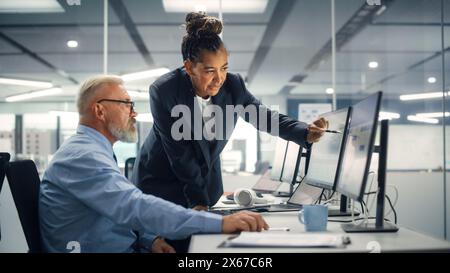 Senior Manager che parla con un uomo d'affari esperto e sicuro di sé mentre utilizza il computer in ufficio moderno. I colleghi discutono di progetti commerciali, finanziari e di marketing. Gli specialisti lavorano in team diversificati. Foto Stock