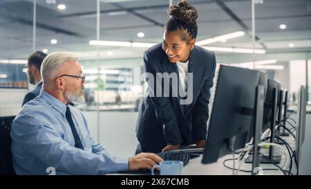 Senior Manager che parla con un uomo d'affari esperto e sicuro di sé mentre utilizza il computer in ufficio moderno. I colleghi discutono di progetti commerciali, finanziari e di marketing. Gli specialisti lavorano in team diversificati. Foto Stock