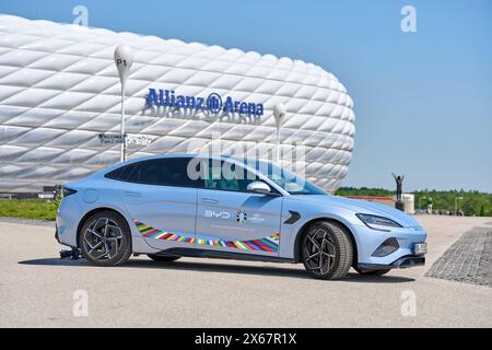 Monaco di Baviera, Germania - 13 maggio 2024: E-car BYD con logo UEFA di fronte allo stadio Allianz Arena di Monaco. Partner ufficiale per la mobilità elettronica di UEFA Euro 2024 *** BYD e-Auto mit UEFA Logo vor dem Fußball Stadion der Allianz Arena di München. Offizieller e-Mobilitätspartner der UEFA Euro 2024 Foto Stock