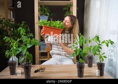 La donna ammira il giardino di erbe in vaso nel suo spazio interno ben illuminato, circondato da piante e attrezzi da giardinaggio su un tavolo di legno. La giardiniera annusa il basilico verde fresco, chiudendo gli occhi con piacere. Foto Stock