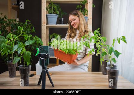 La creatrice di contenuti sorride mentre registra un video in cui si tiene una grande pentola di basilico, circondata dal suo fiorente giardino interno di varie piante in vaso. Foto Stock