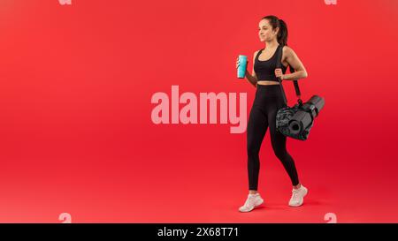 Donna attiva in abbigliamento sportivo che trasporta attrezzature da palestra su sfondo rosso Foto Stock