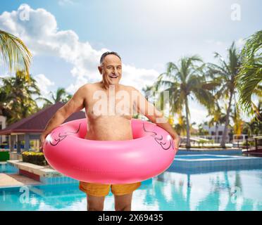 Uomo maturo felice che indossa pantaloncini da nuoto e anello gonfiabile in gomma flamingo vicino a una piscina Foto Stock